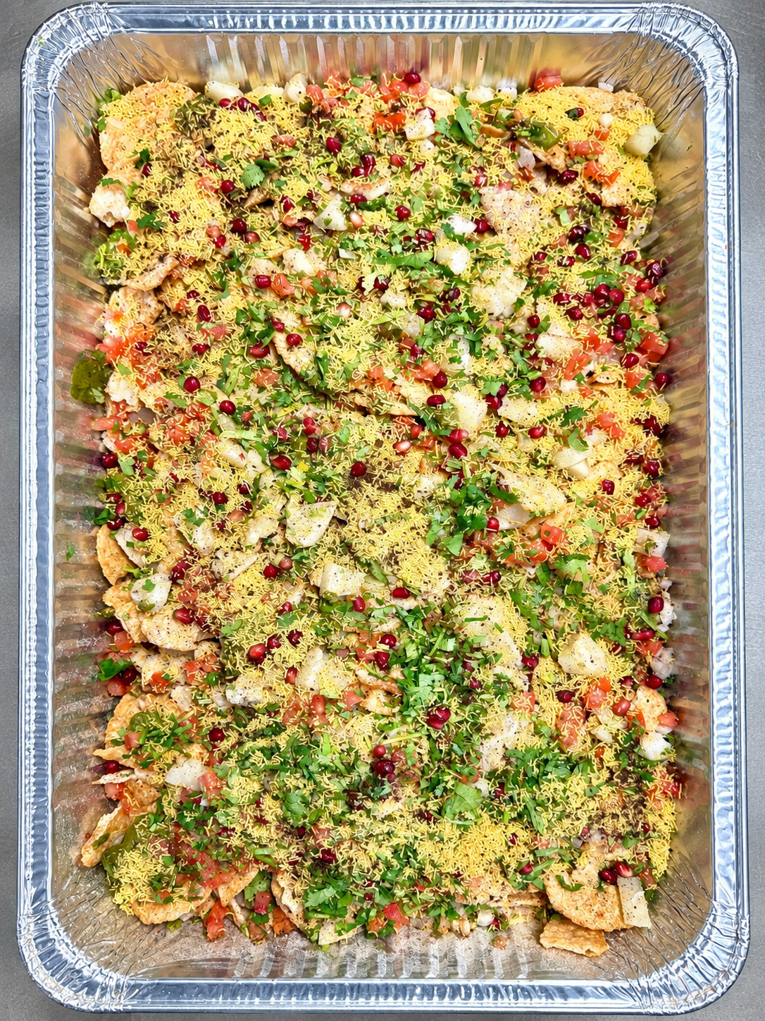 A party-size catering tray of chaat topped with sev, pomegranate and cilantro